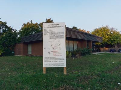 A public notice sign for the City of Hamilton stands in a grassy area in front of a single-story brick building with a dark, sloped roof.