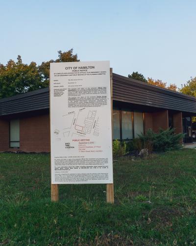 A public notice sign for the City of Hamilton stands in a grassy area in front of a single-story brick building with a dark, sloped roof.