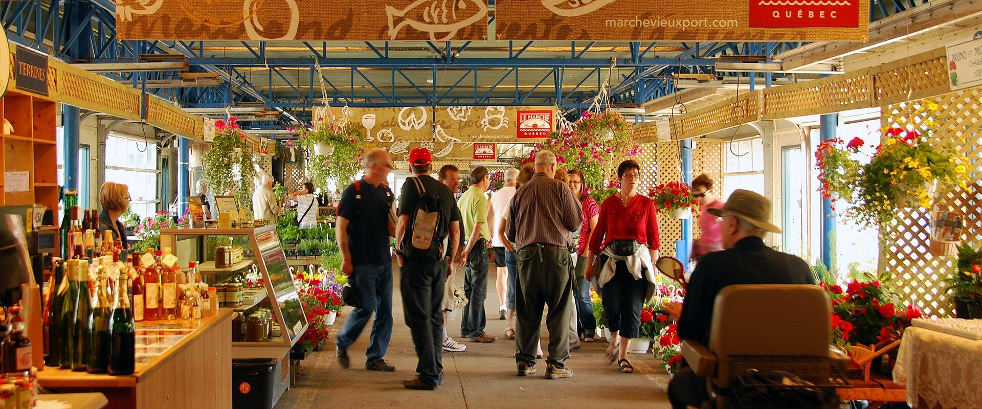 A customer in a hat stands at a vendor's counter inside a market, looking at cured sausages in a refrigerated display case, with a red sign hanging above reading "LE MARCHÉ DU VIEUX-PORT QUÉBEC".