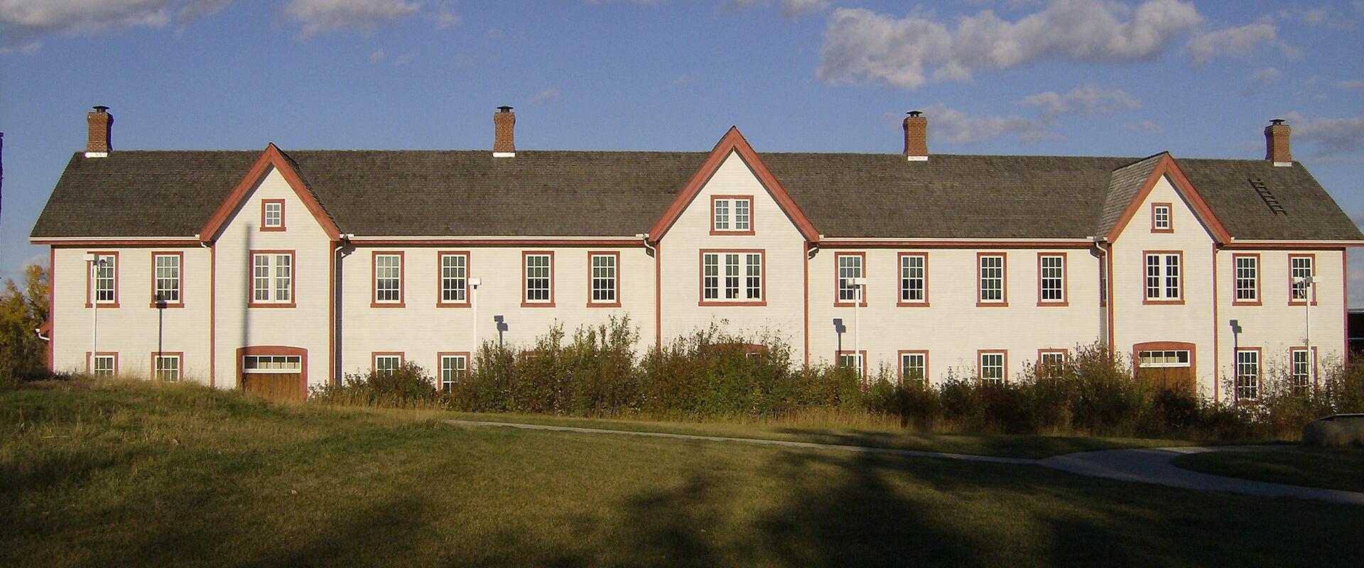 A long, two-story white building with red trim and a grey shingled roof, featuring multiple chimneys and symmetrically arranged windows, set against a blue sky with scattered clouds and a grassy foreground.