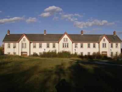 A long, two-story white building with red trim and a grey shingled roof, featuring multiple chimneys and symmetrically arranged windows, set against a blue sky with scattered clouds and a grassy foreground.