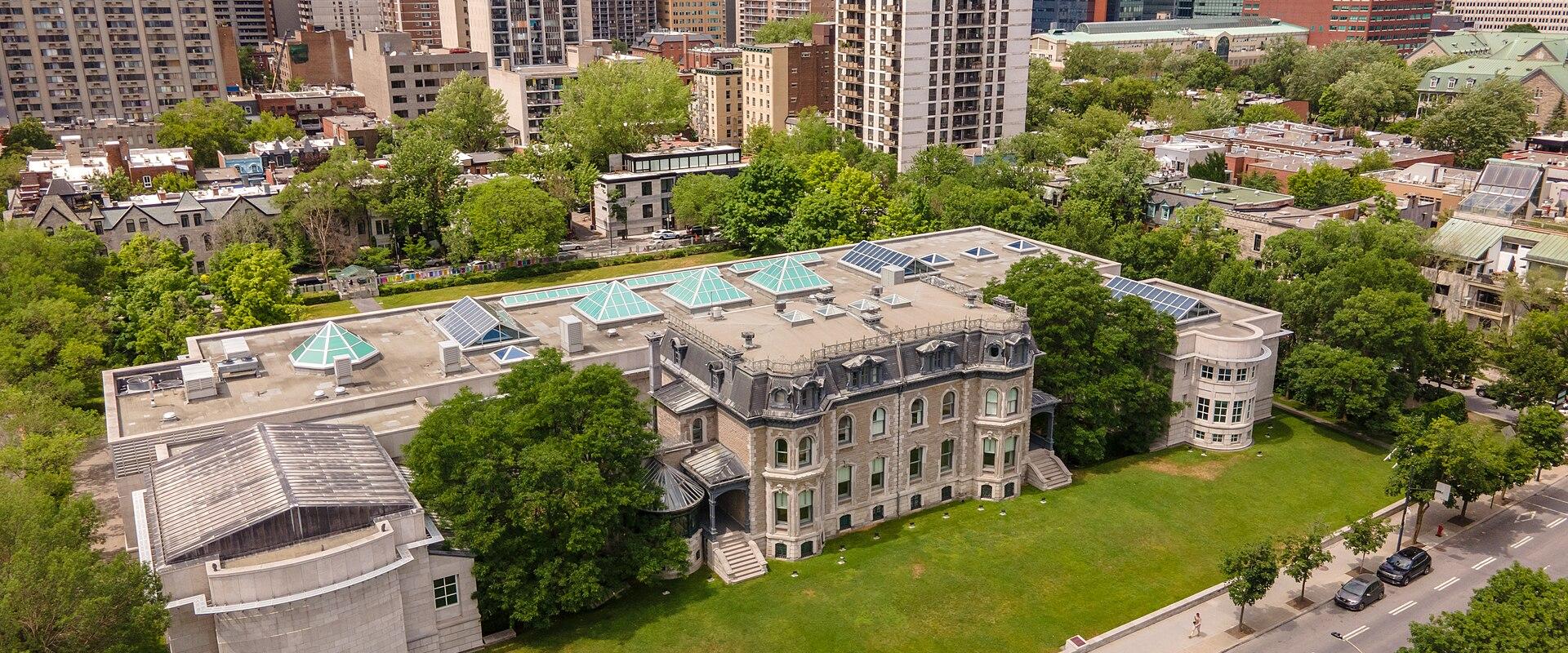 An aerial view of a large, historic stone mansion with a modern concrete annex and a roof featuring multiple glass skylights, situated on a green lawn next to a city street with high-rise buildings in the background.