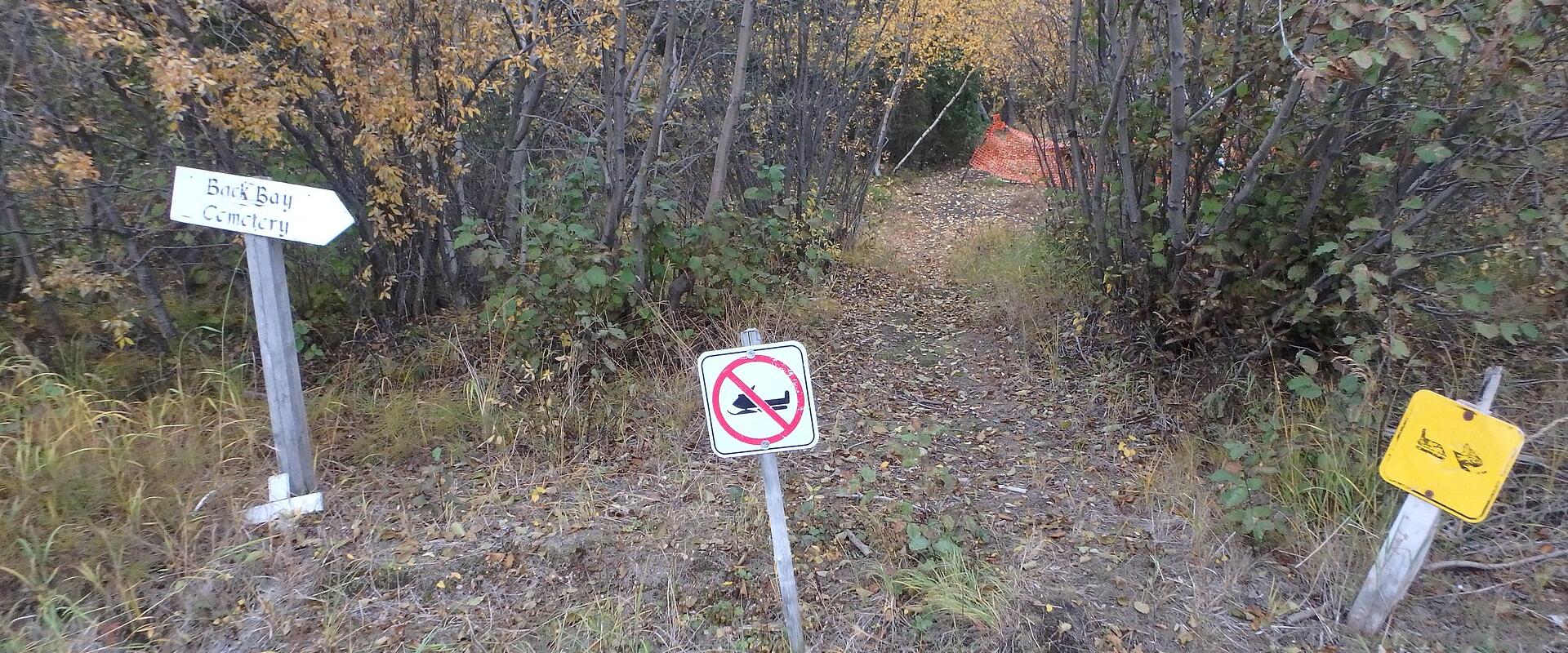 A dirt path through a wooded area with autumn foliage, featuring a white directional sign pointing left that reads "Back Bay Cemetery," a "no vehicles" prohibition sign, and a yellow warning sign on the right.