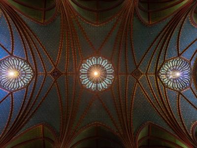 An upward view of an ornate, vaulted ceiling featuring blue and gold star patterns, ribbed arches, and three circular stained-glass windows illuminated by artificial light.