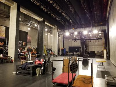 A backstage or loading dock area of a performance venue, showing instrument cases, carts, and stage rigging.