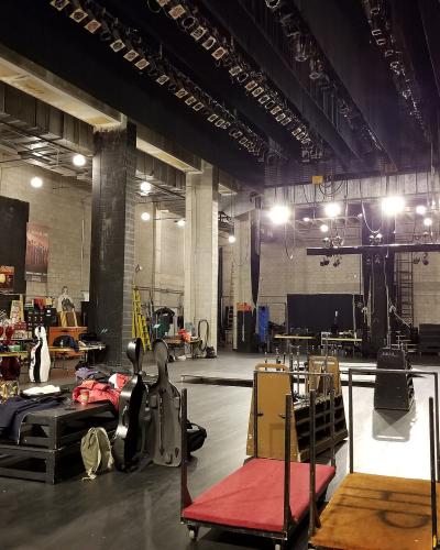 A backstage or loading dock area of a performance venue, showing instrument cases, carts, and stage rigging.