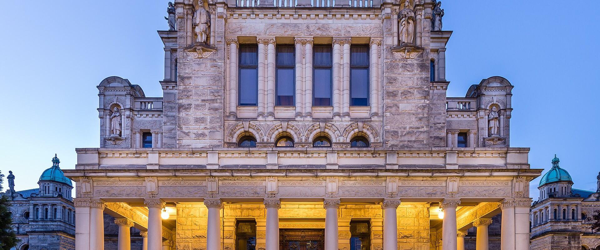 A grand, ornate stone building with a central portico supported by large columns, illuminated at twilight against a blue sky, featuring statues in niches and green-domed towers on the flanking wings.