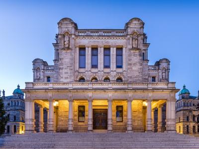 A grand, ornate stone building with a central portico supported by large columns, illuminated at twilight against a blue sky, featuring statues in niches and green-domed towers on the flanking wings.