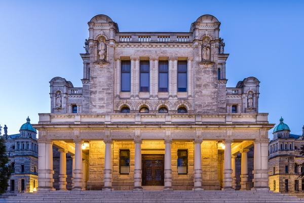 A grand, ornate stone building with a central portico supported by large columns, illuminated at twilight against a blue sky, featuring statues in niches and green-domed towers on the flanking wings.