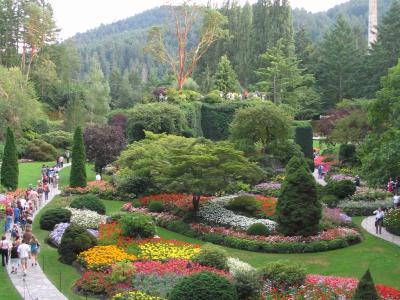 A high-angle view of a terraced botanical garden featuring winding stone pathways, vibrant flowerbeds in yellow, red, and white, manicured hedges, and a tall brick chimney visible in the background forest.