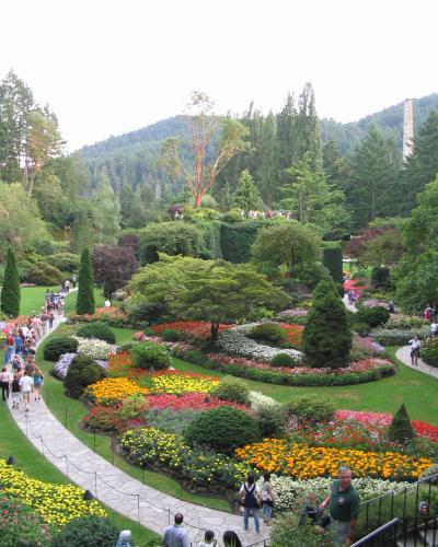 A high-angle view of a terraced botanical garden featuring winding stone pathways, vibrant flowerbeds in yellow, red, and white, manicured hedges, and a tall brick chimney visible in the background forest.