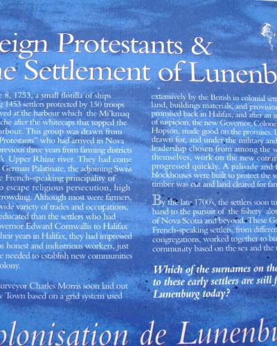 A close-up photograph of a blue informational sign titled "Foreign Protestants & the Settlement of Lunenburg" with a logo for the Town of Lunenburg in the top right corner.