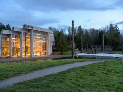 A modern concrete building with large glass windows displaying interior artifacts, situated next to a grassy area with a path and several tall wooden totem poles in the background.