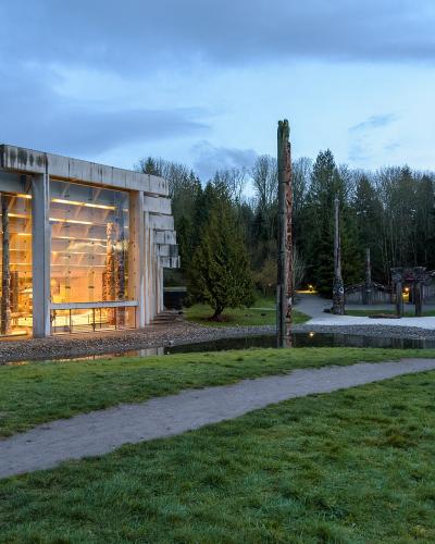 A modern concrete building with large glass windows displaying interior artifacts, situated next to a grassy area with a path and several tall wooden totem poles in the background.