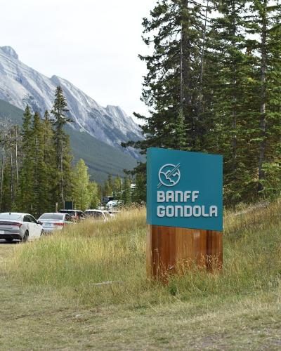 A teal and wood sign reading "BANFF GONDOLA" stands in a grassy area near a parking lot with cars, with a mountain slope and evergreen trees in the background.