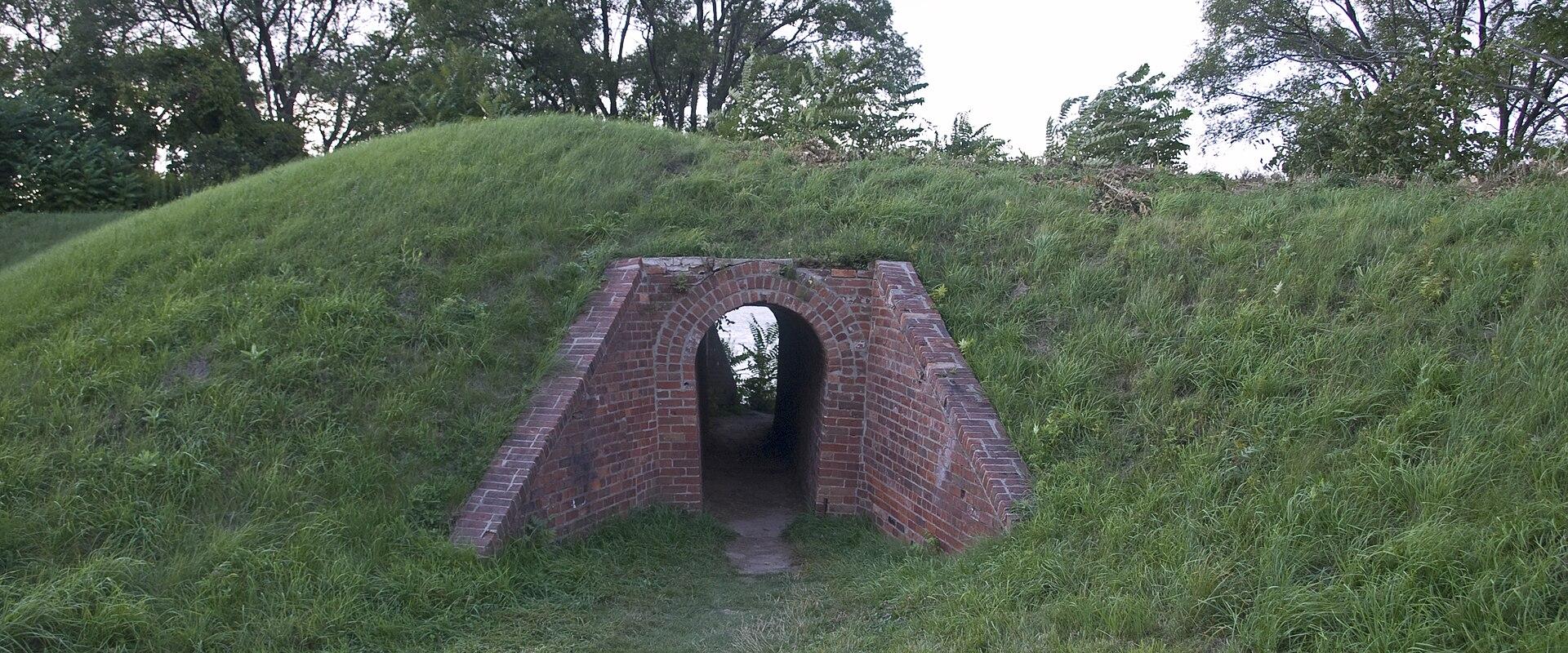 A grassy earthen mound with a red brick arched tunnel entrance set into its side, surrounded by trees.