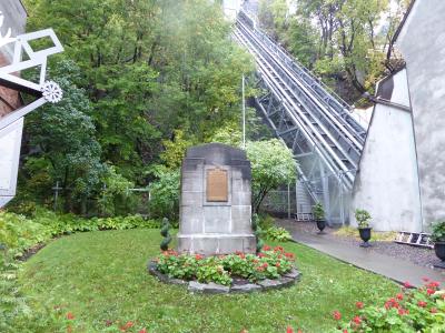 A stone monument with a bronze plaque stands in a grassy garden area, with a large inclined funicular railway structure rising up a wooded hillside in the background.