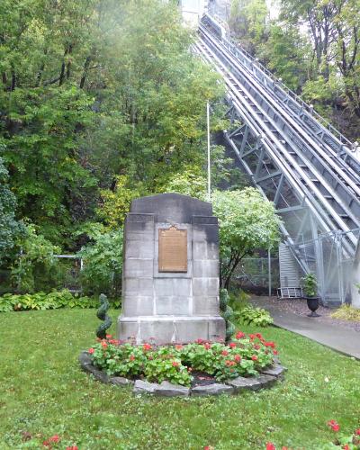 A stone monument with a bronze plaque stands in a grassy garden area, with a large inclined funicular railway structure rising up a wooded hillside in the background.