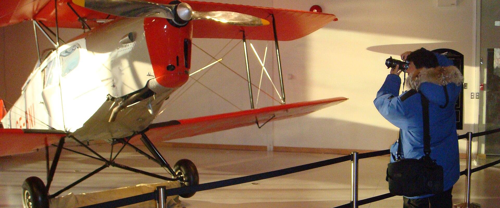 An indoor museum exhibit featuring a vintage red and white biplane on display, with a visitor in winter clothing photographing it.