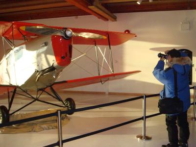 An indoor museum exhibit featuring a vintage red and white biplane on display, with a visitor in winter clothing photographing it.