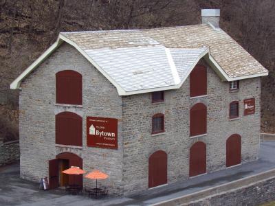 A large stone building with a shingled roof, featuring a prominent brown sign on the facade that reads "musée Bytown museum" and "Ottawa commence ici... where Ottawa begins," along with orange umbrellas outside.