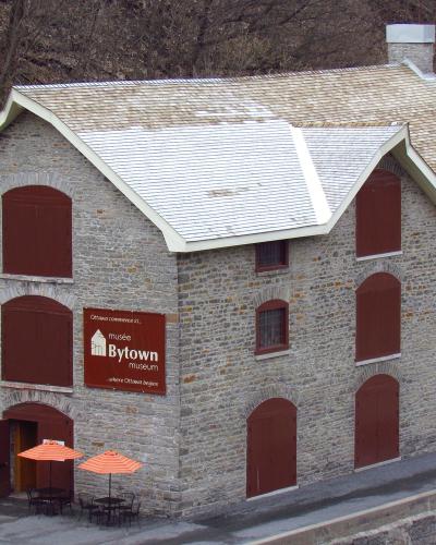A large stone building with a shingled roof, featuring a prominent brown sign on the facade that reads "musée Bytown museum" and "Ottawa commence ici... where Ottawa begins," along with orange umbrellas outside.