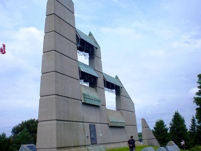 A low-angle photograph of a large, modern concrete bell tower structure with a distinctive stepped, angular design and green copper roofs over the belfry openings, situated on a grassy hill with a Canadian flag visible on the left.