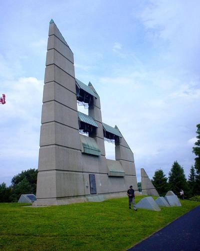 A low-angle photograph of a large, modern concrete bell tower structure with a distinctive stepped, angular design and green copper roofs over the belfry openings, situated on a grassy hill with a Canadian flag visible on the left.