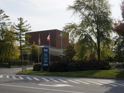 A street-level view of a modern brick building complex surrounded by trees, featuring a prominent blue sign listing "Shaw Festival," "Festival Theatre," "Jackie Maxwell Studio," and "Shaw Theatre," with Canadian and other flags flying nearby.
