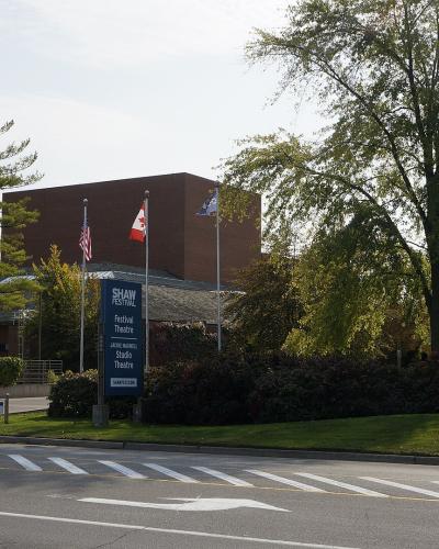 A street-level view of a modern brick building complex surrounded by trees, featuring a prominent blue sign listing "Shaw Festival," "Festival Theatre," "Jackie Maxwell Studio," and "Shaw Theatre," with Canadian and other flags flying nearby.