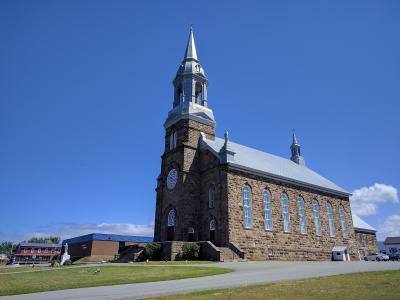 A large stone church with a prominent central steeple and a smaller rear steeple, set against a blue sky with a road and other buildings in the foreground.