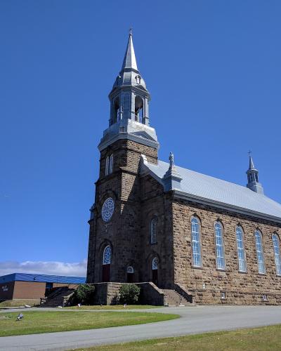 A large stone church with a prominent central steeple and a smaller rear steeple, set against a blue sky with a road and other buildings in the foreground.