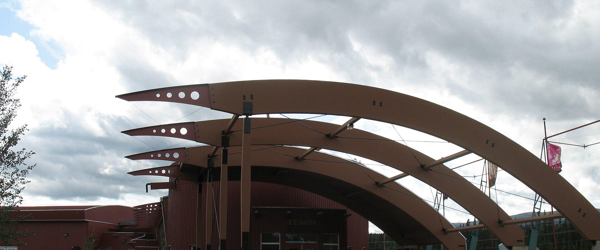 The image shows the entrance to a modern building with a distinctive reddish-brown facade and a large, sculptural canopy made of curved, layered beams, with the word "YUKON" visible above the main doors.