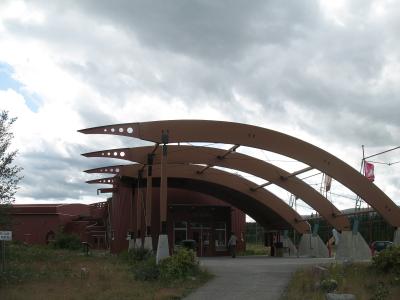 The image shows the entrance to a modern building with a distinctive reddish-brown facade and a large, sculptural canopy made of curved, layered beams, with the word "YUKON" visible above the main doors.