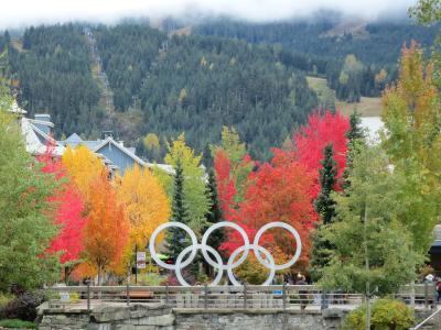 A large white Olympic rings sculpture stands in a plaza surrounded by autumn-colored trees, with ski lifts visible on a forested mountain in the background.