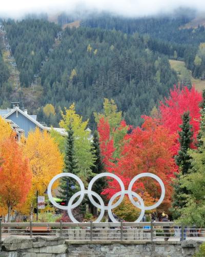 A large white Olympic rings sculpture stands in a plaza surrounded by autumn-colored trees, with ski lifts visible on a forested mountain in the background.