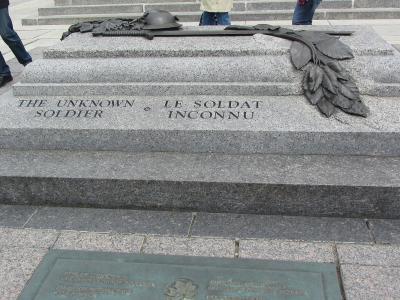A close-up view of a granite tombstone inscribed with "THE UNKNOWN SOLDIER" and "LE SOLDAT INCONNU," featuring bronze artifacts including a helmet, rifle, and maple leaf branch, situated on a paved plaza.