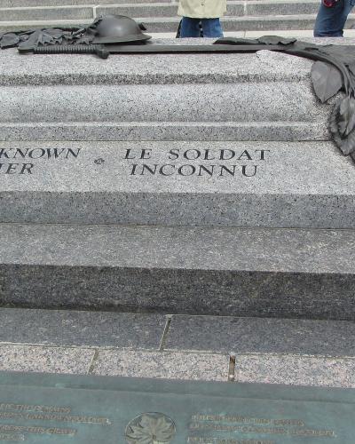 A close-up view of a granite tombstone inscribed with "THE UNKNOWN SOLDIER" and "LE SOLDAT INCONNU," featuring bronze artifacts including a helmet, rifle, and maple leaf branch, situated on a paved plaza.