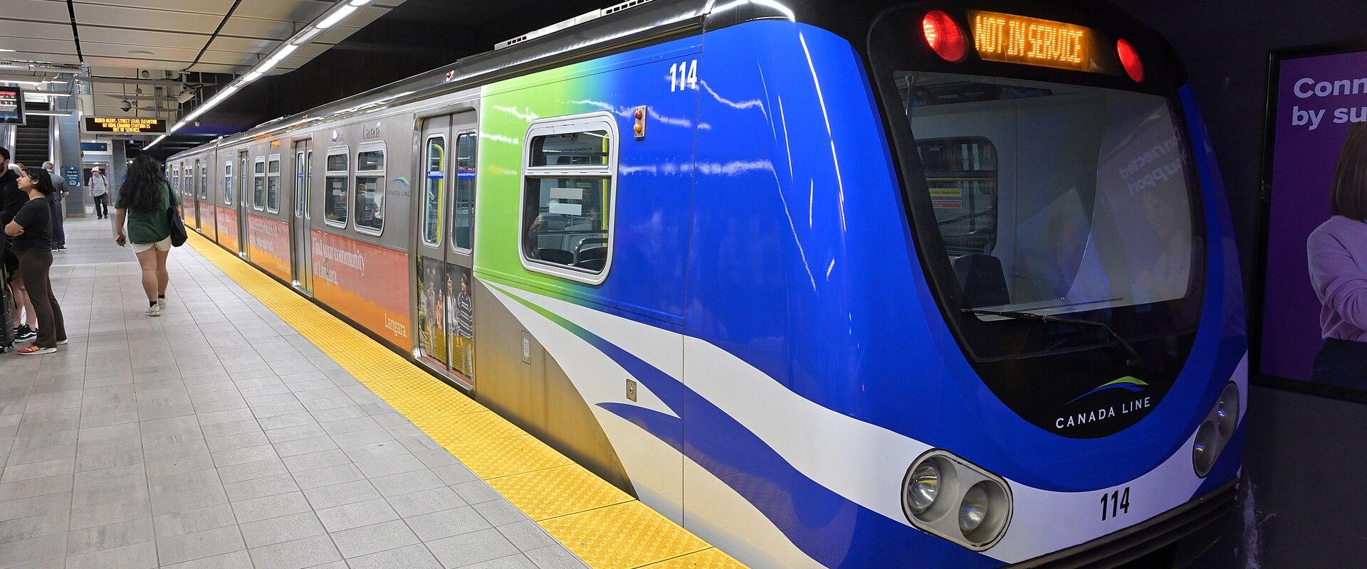 A blue, green, and silver SkyTrain train (number 114) stopped at an underground station platform with passengers waiting.