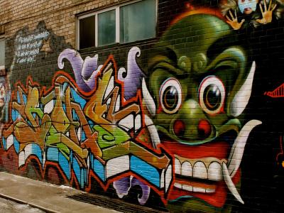 A close-up view of a brick wall covered in colorful street art, featuring a large green dragon-like creature with tusks and a stylized graffiti tag in the foreground.