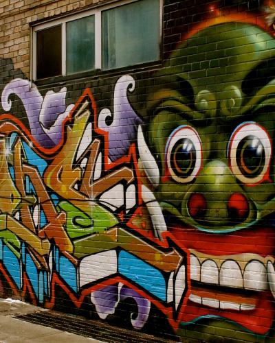 A close-up view of a brick wall covered in colorful street art, featuring a large green dragon-like creature with tusks and a stylized graffiti tag in the foreground.