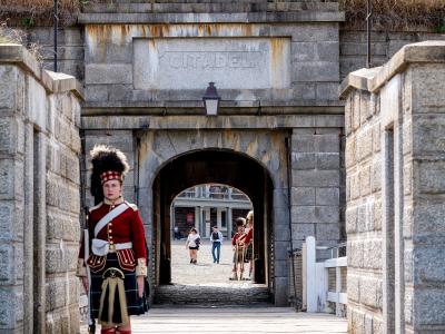 A stone gateway with the word "CITADEL" carved into the keystone, flanked by stone walls and a grassy rampart, with a soldier in a red coat and kilt standing guard in the foreground.
