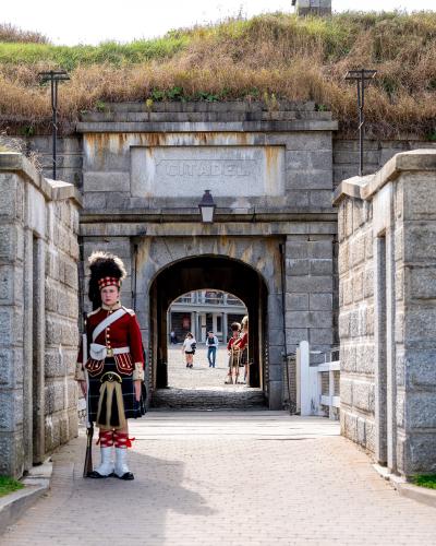 A stone gateway with the word "CITADEL" carved into the keystone, flanked by stone walls and a grassy rampart, with a soldier in a red coat and kilt standing guard in the foreground.