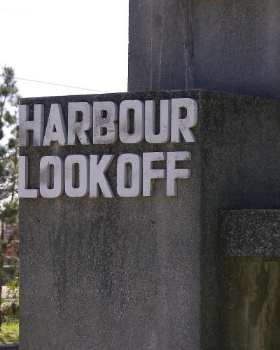 A close-up of a concrete structure featuring raised white lettering that reads "HARBOUR LOOKOFF", with pine trees and a fence visible in the background.