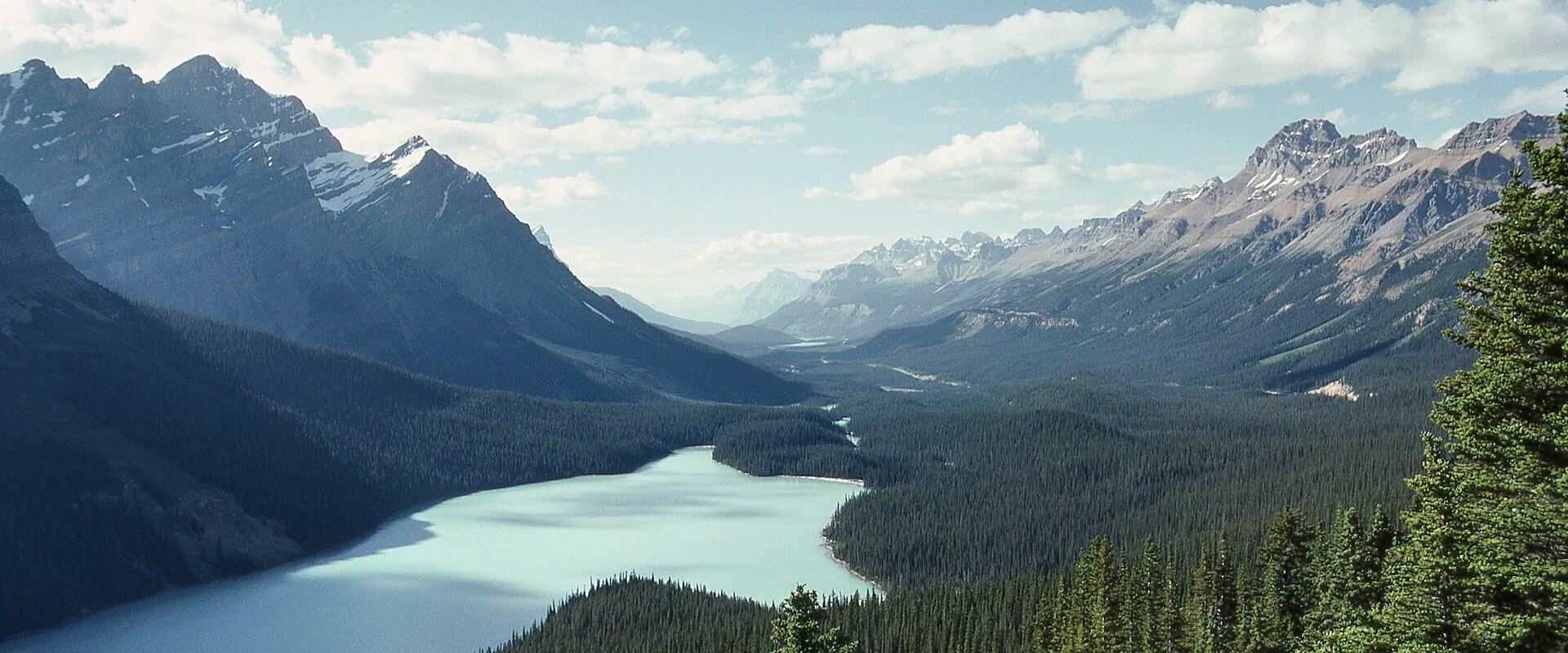 A high-angle landscape photograph showing a turquoise glacial lake winding through a valley of dense coniferous forest, surrounded by rugged, rocky mountains under a partly cloudy sky.
