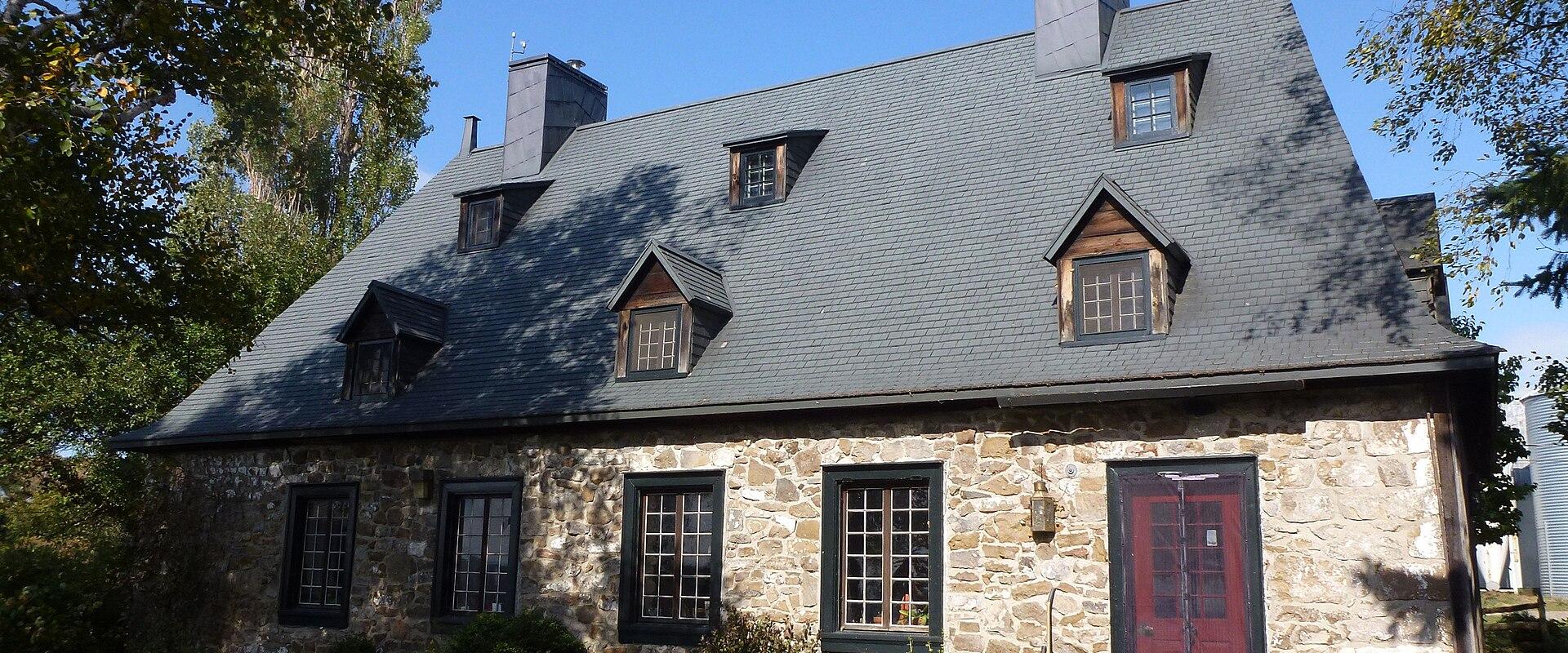 A two-story stone house with a steep slate roof, dormer windows, black shutters, and a red door, set against a blue sky with trees and a lawn.