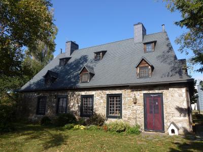 A two-story stone house with a steep slate roof, dormer windows, black shutters, and a red door, set against a blue sky with trees and a lawn.