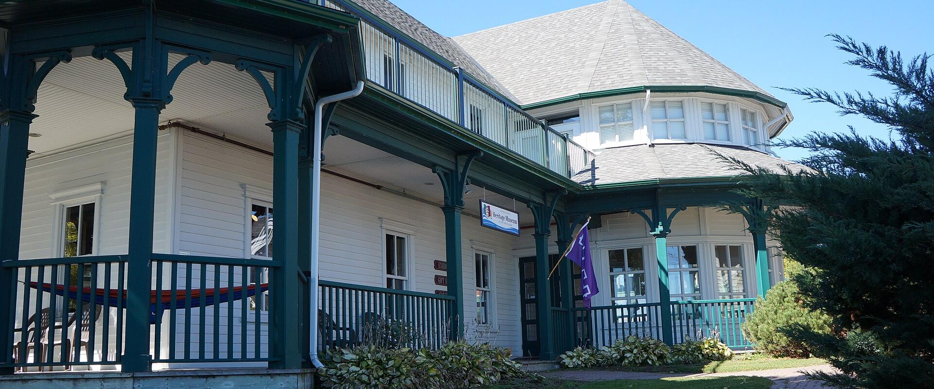 A white wooden building with green trim, featuring a wraparound porch, a balcony, and a distinctive octagonal turret, with a blue sign hanging from the porch.