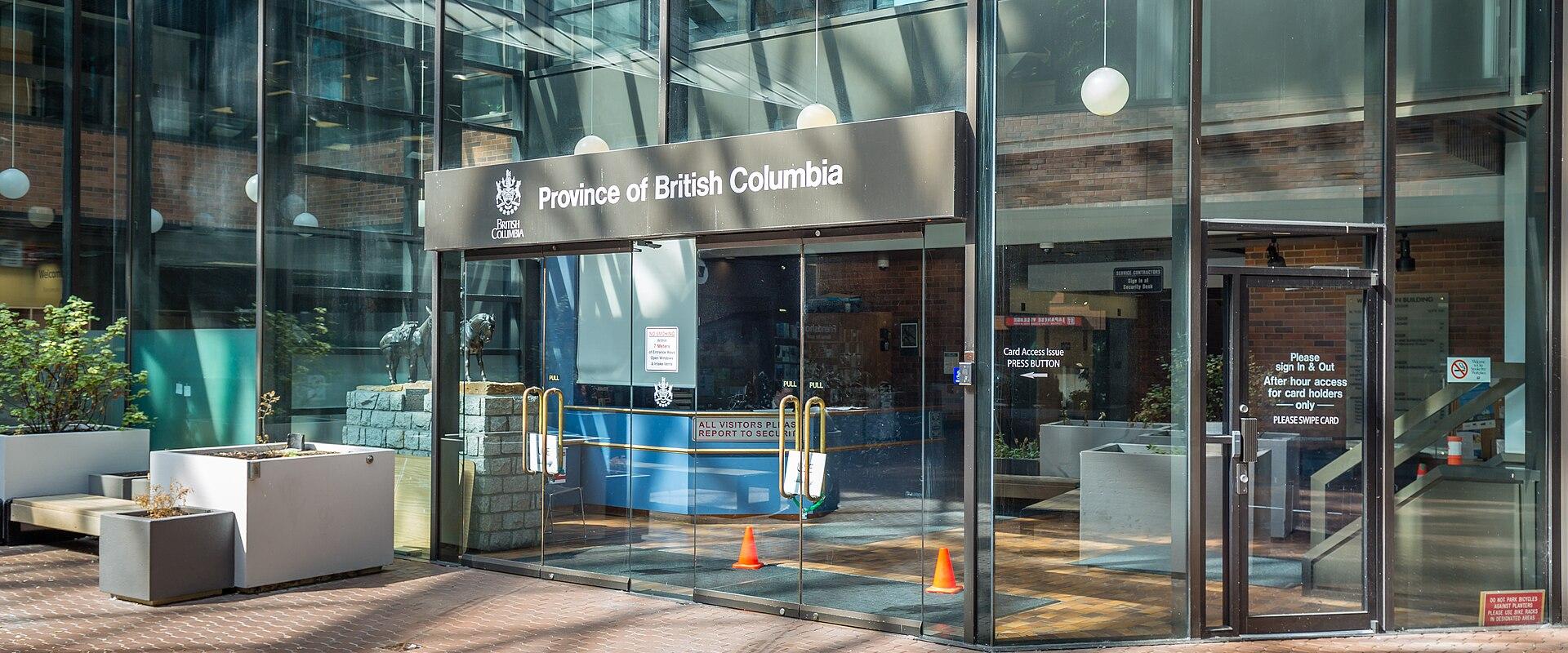 The image shows the glass entrance to a modern building with a sign reading "Province of British Columbia" above the doors, located within a paved courtyard area.