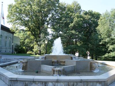 A multi-tiered stone fountain with water jets, situated on a paved terrace with a large stone building and a flagpole visible to the left and dense trees in the background.